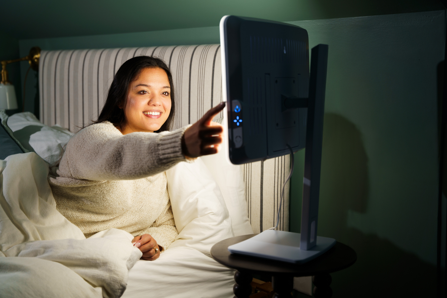 A woman turning on a light therapy lamp next to her bed