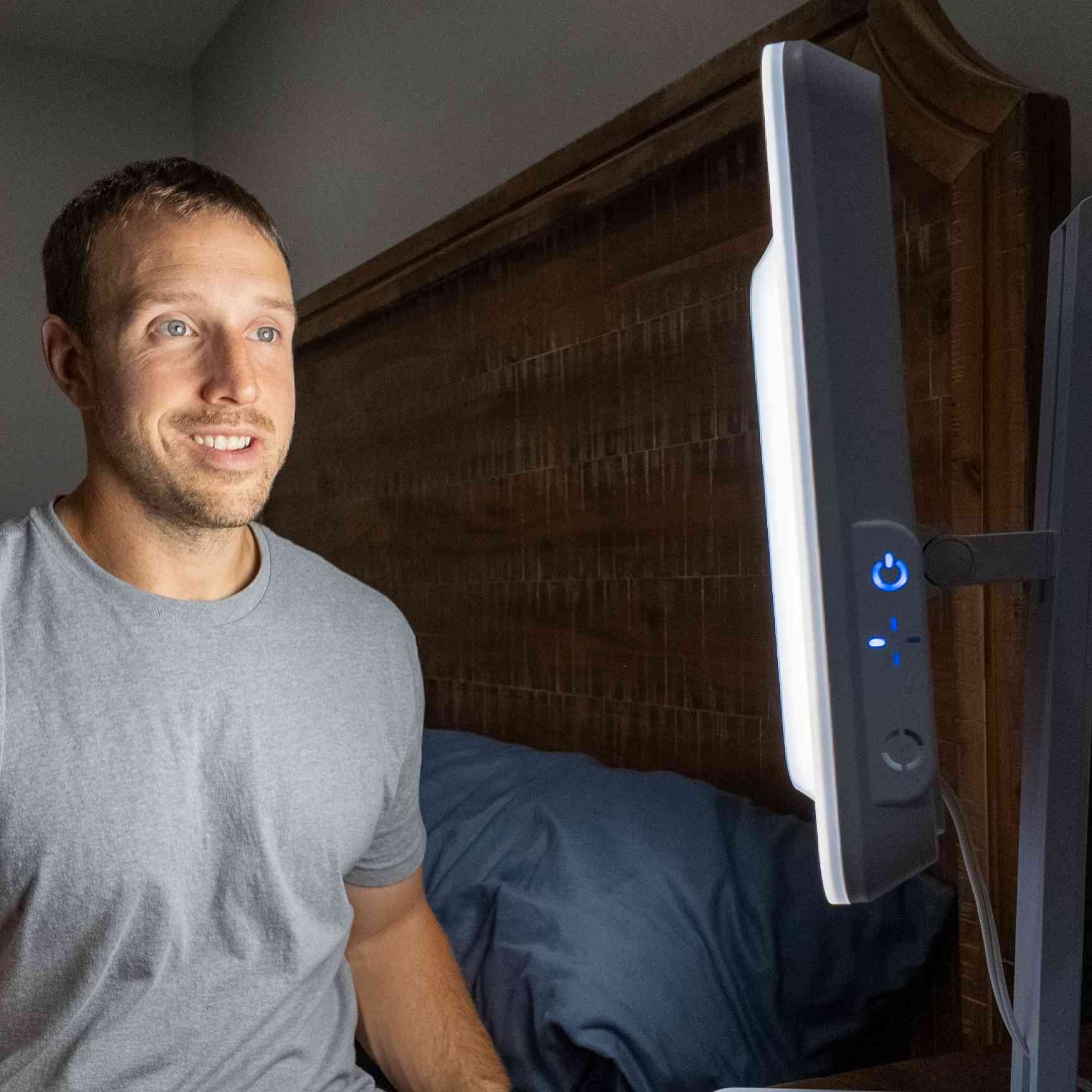 A man sitting on his bed in front of a white light therapy lamp