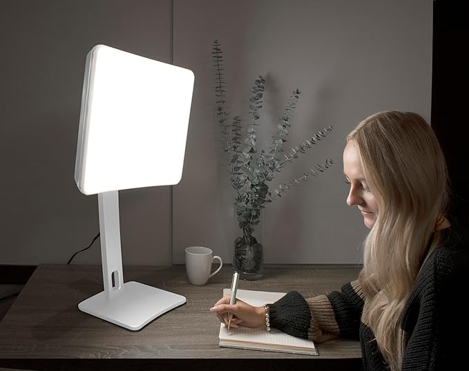 A woman sitting next to a white light therapy lamp while writing