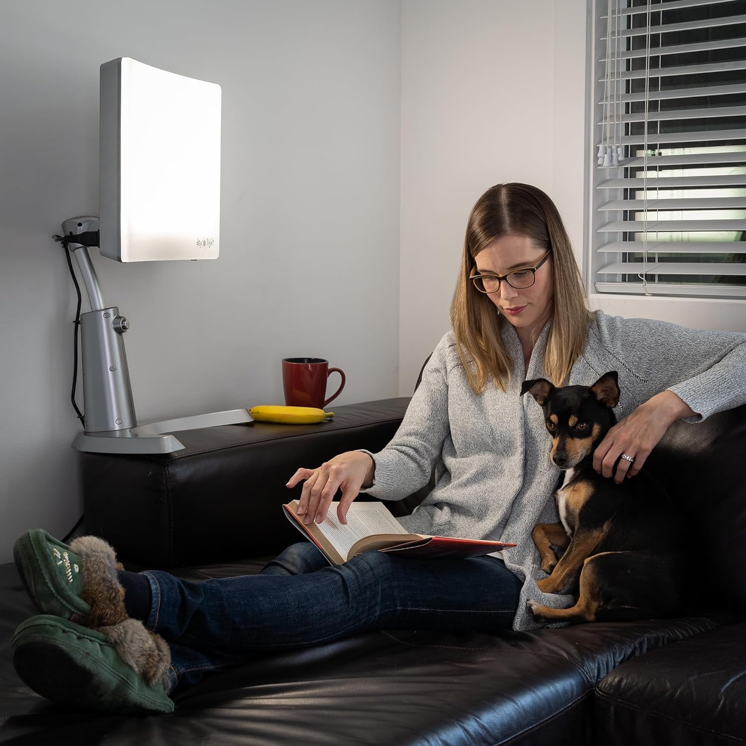 A woman sitting on a couch with her dog reading. A therapy lamp on the arm of the couch.