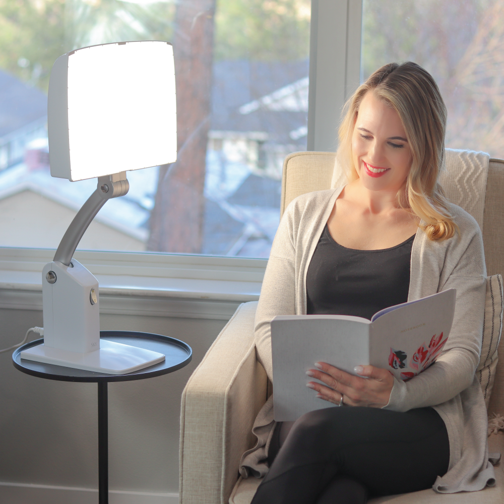 A woman reading a book sitting next to a white therapy lamp