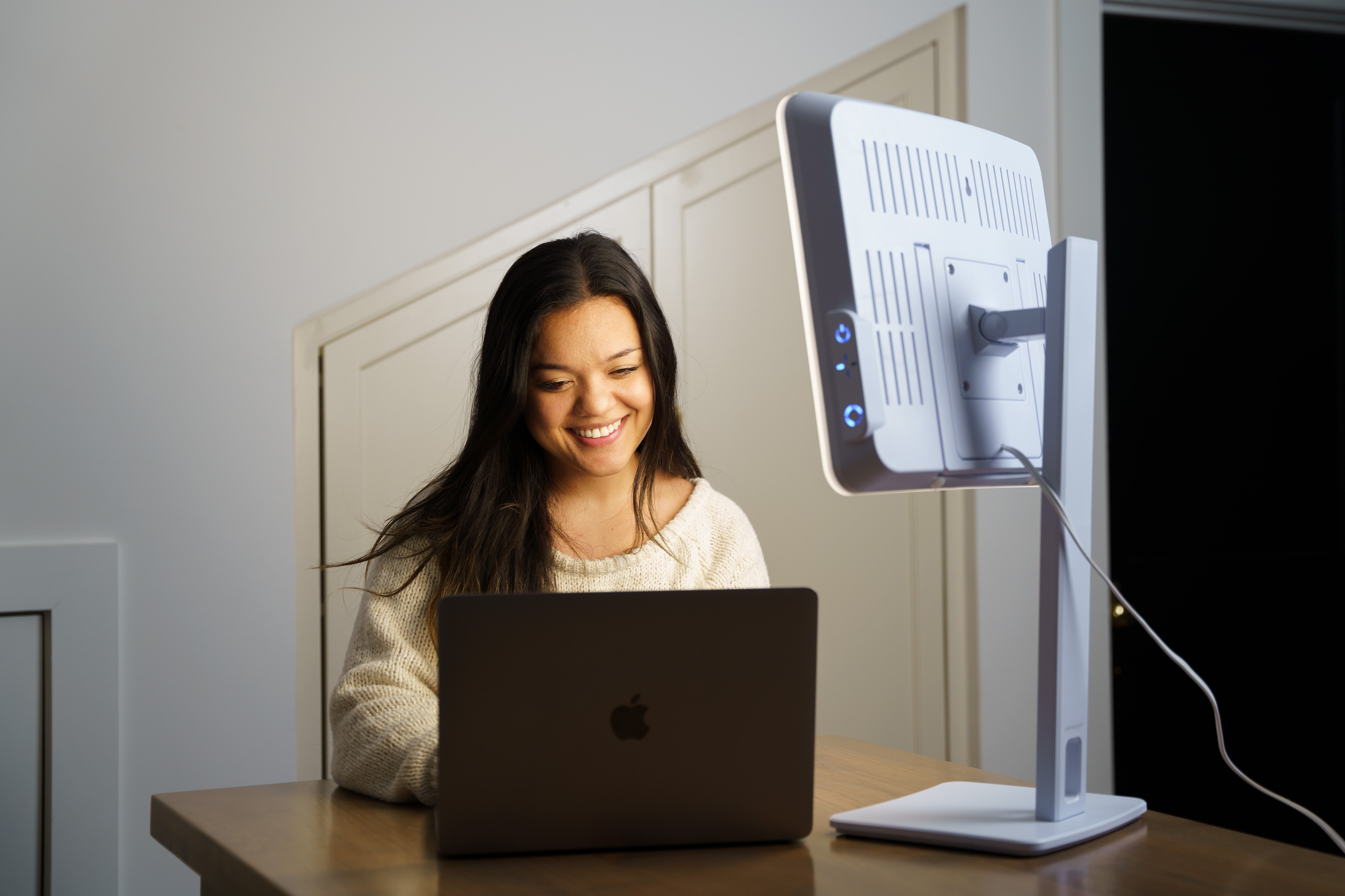 A smiling woman on a laptop in front of a therapy lamp